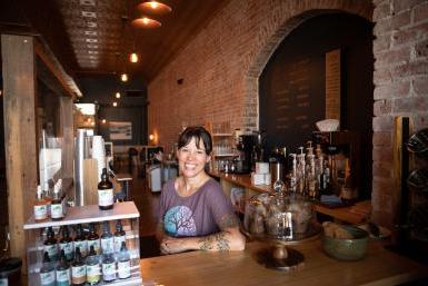 Artisan Mark owner Elayne Woods Jones stands behind the coffeeshop's counter. 在她身后, 很长一段, exposed brick wall extends to seating at the back of the shop, 头顶温暖的灯光.